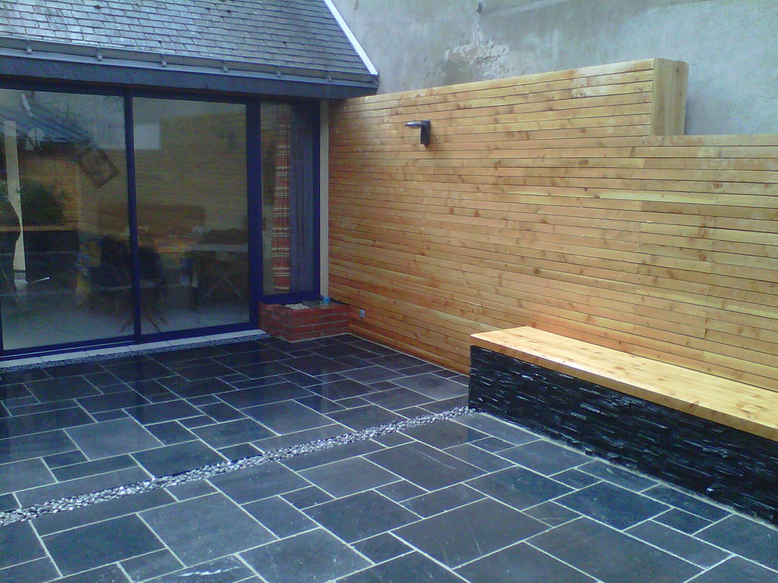 Terrasse en carrelage avec séparation décorative en graviers, un mur et banc en bois et une baie vitrée par Aux 1001 Saisons